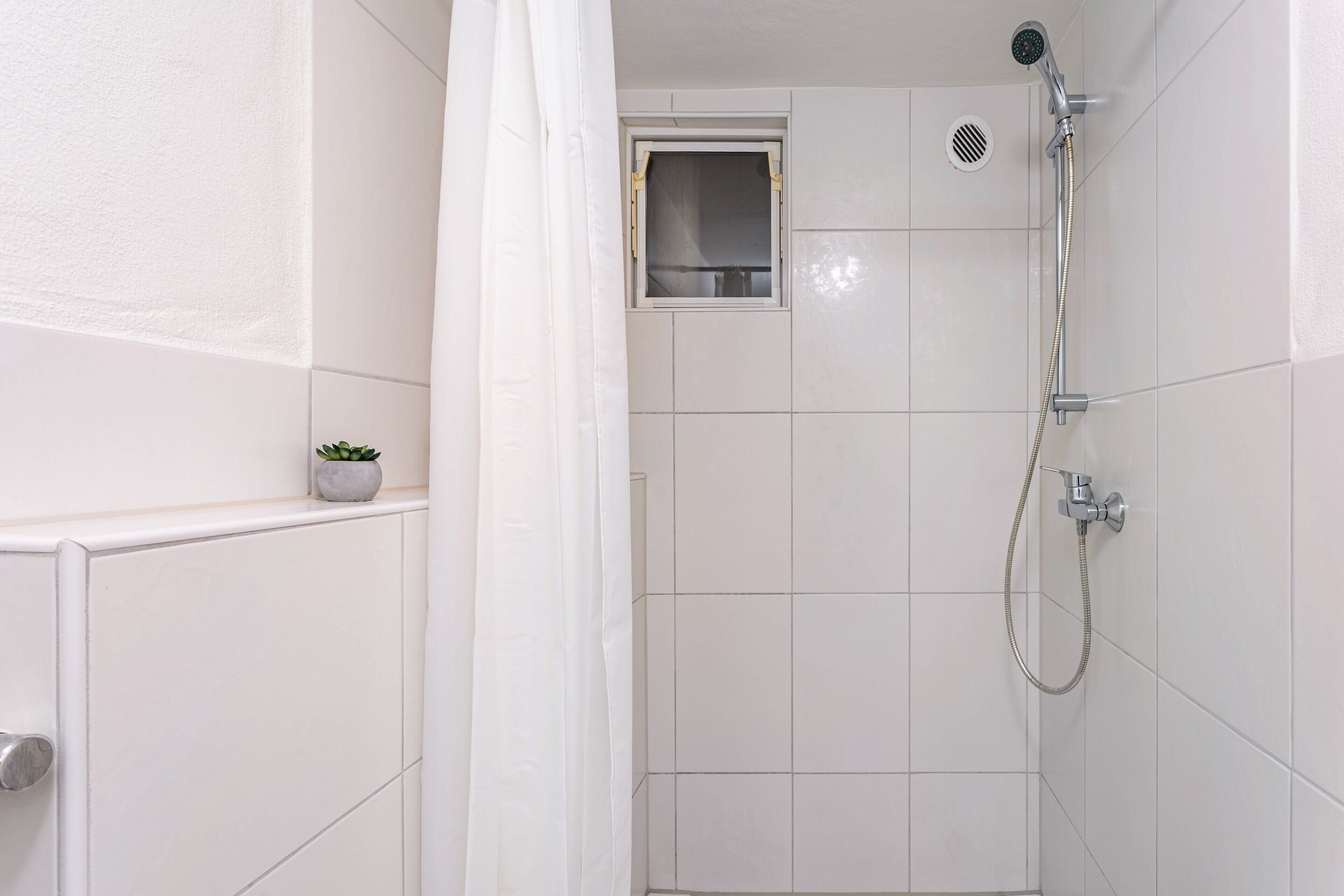 Ein Badezimmer mit weißen Fliesen, einer Dusche mit Duschbrause, einem kleinen Fenster und einem Topf mit einer Pflanze auf dem Heizkörper.