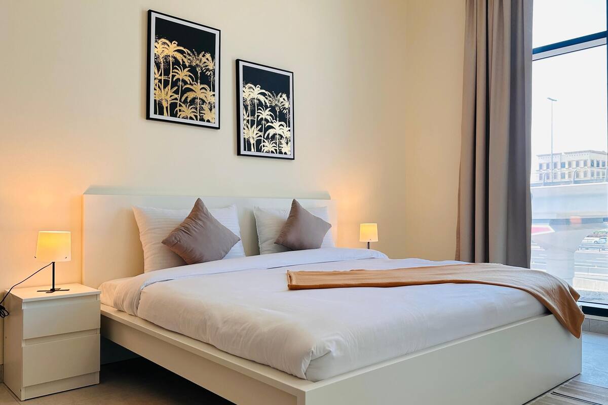 A modern hotel room with a large bed, two bedside tables with lamps, and a view of the city through large windows.