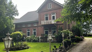 A large brick house with a balcony, surrounded by a well-maintained garden with various plants and a gravel driveway.