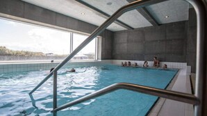 Una piscina interior con agua azul cristalina, una escalera metálica y vista al cielo a través de amplios ventanales.
