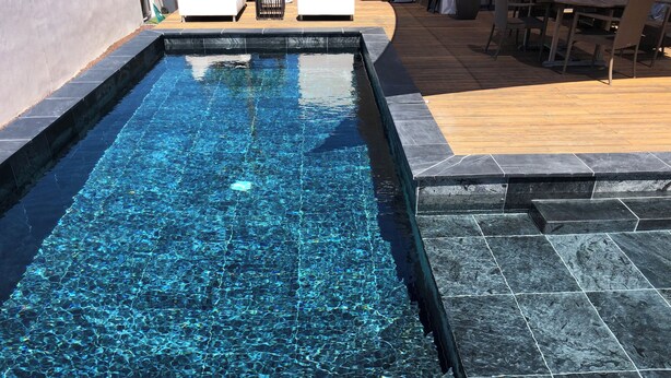 A rectangular swimming pool with clear blue water, surrounded by dark stone tiles and wooden decking.