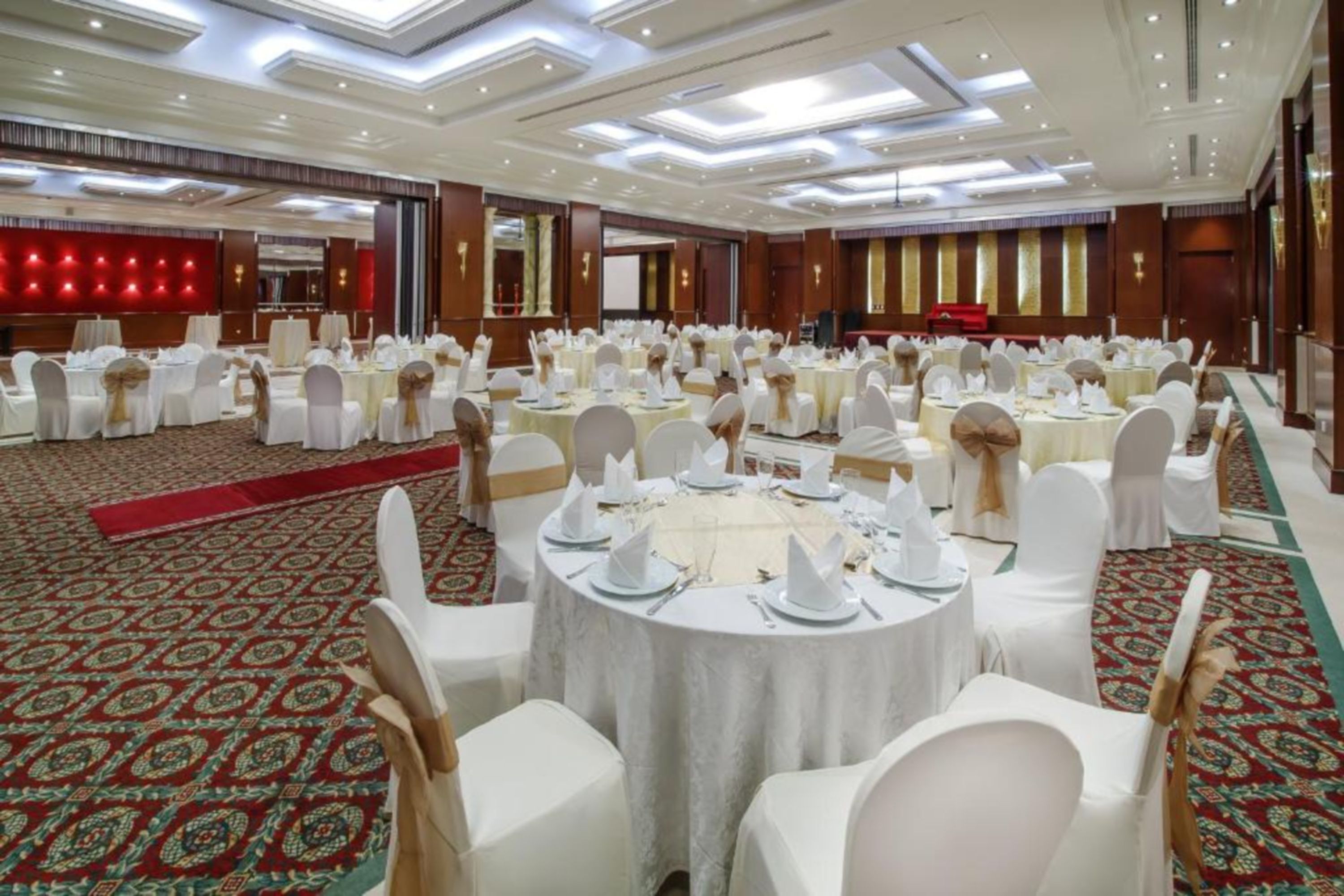 A banquet hall with round tables set for dining, white chairs, and a red carpet.