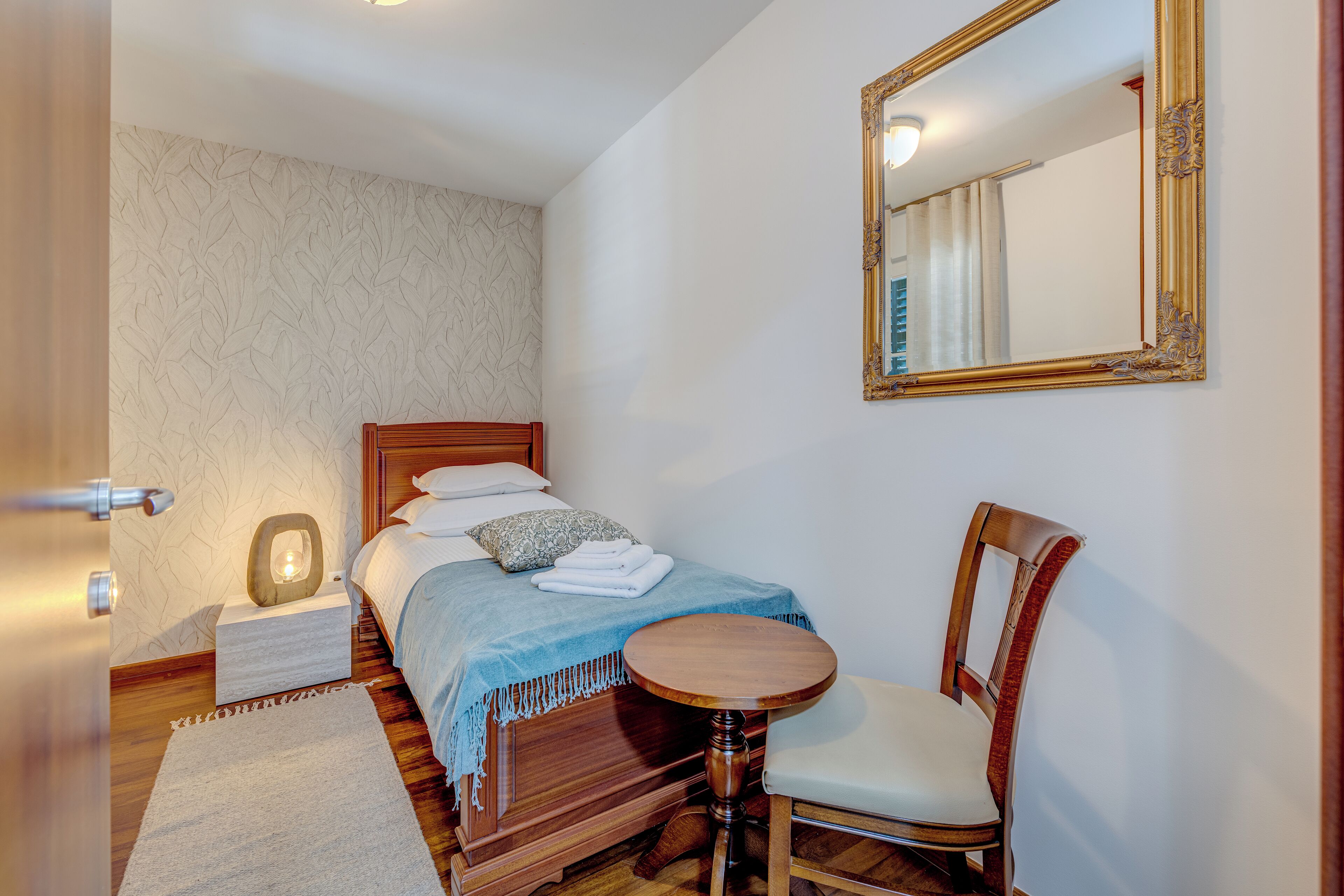 A bedroom with a bed, a wooden chair, a round table, a mirror, and a nightstand.