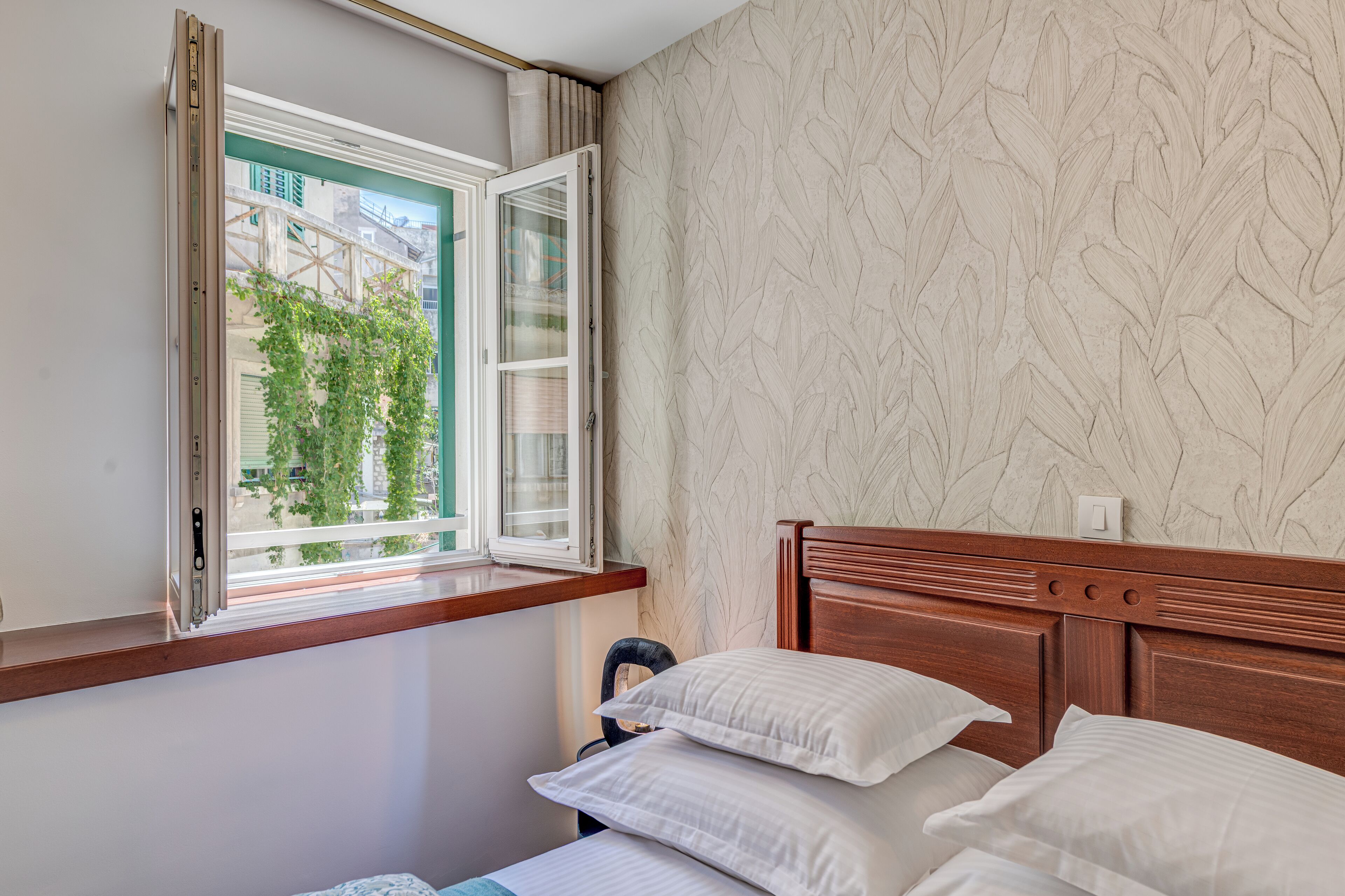 A hotel room with a bed, a window with a view of buildings and trees, and a patterned wall.