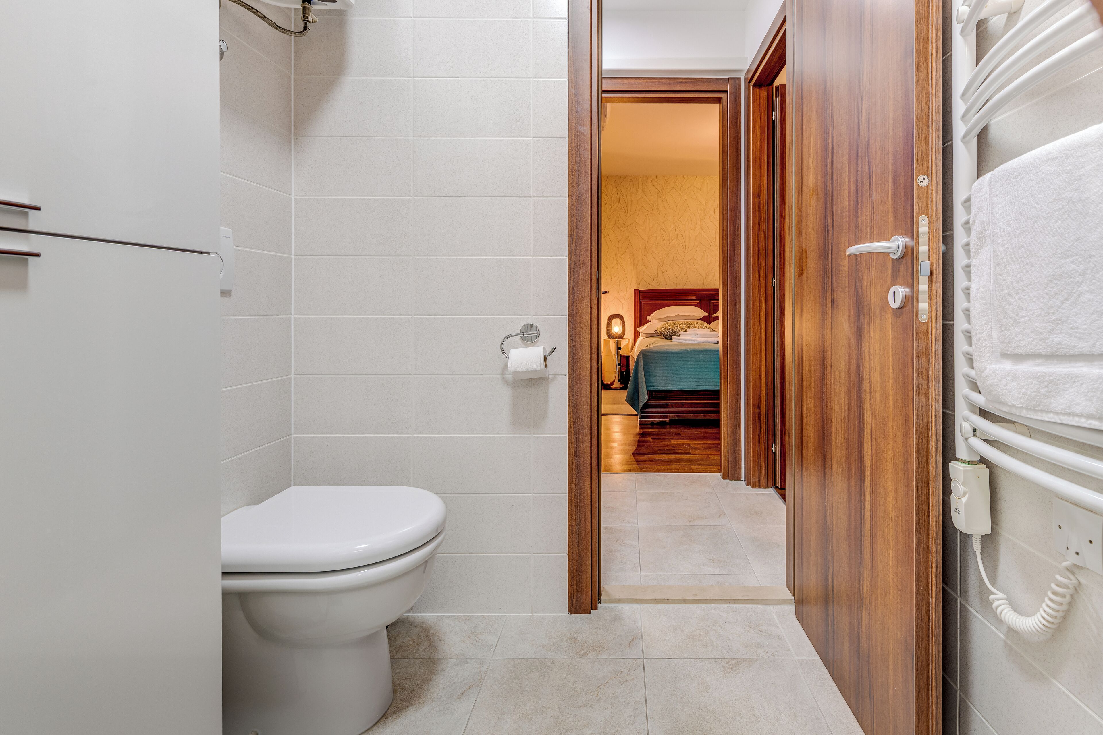 A bathroom with a toilet, a shower area, and a wooden door leading to a bedroom with a bed and a nightstand.