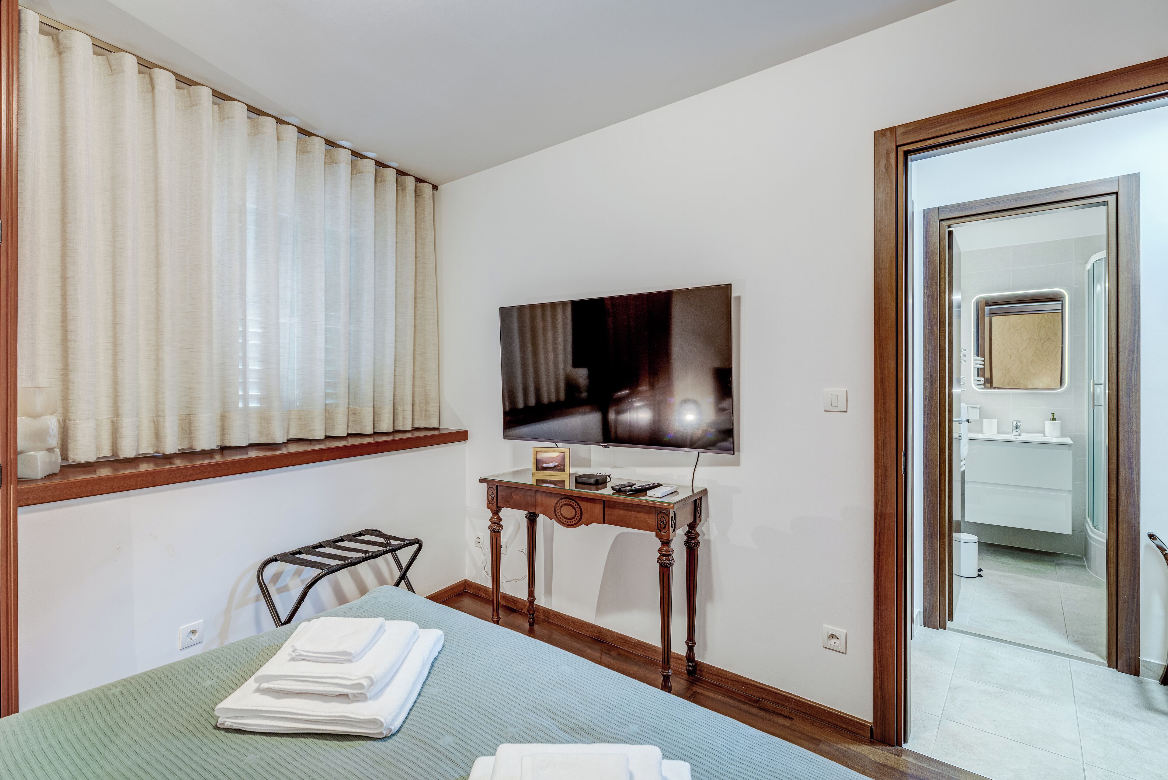 A hotel room with a bed, a desk with a flat-screen TV, a window with curtains, and a bathroom visible through an open door.