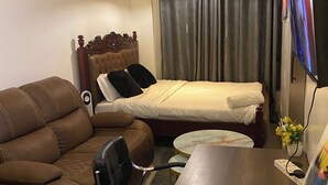 Une chambre d’hôtel avec un canapé en cuir marron, un lit en bois avec une tête de lit, une petite table ronde, un téléviseur et un rideau.