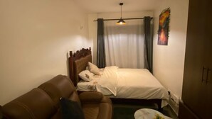 Une chambre d’hôtel avec un canapé en cuir marron, une petite table ronde sur laquelle se trouve un vase, un lit avec du linge de maison blanc, une tête de lit, un luminaire au plafond et un tableau accroché au mur.