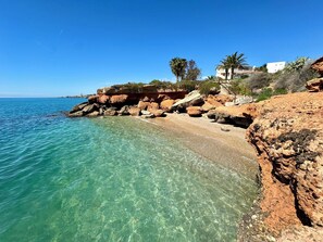 Ein felsiger Strand mit klarem türkisblauem Wasser, Palmen und einem Gebäude mit rotem Dach.
