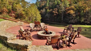 A circular stone fire pit surrounded by wooden Adirondack chairs, set on a red patio with a stone wall and a view of a golf course and forest.