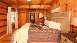 Une chambre dans une cabane en bois, avec un lit, une table de chevet, une lampe, un miroir et une pancarte sur laquelle on peut lire : « l’amour, et avant tout, l’amour ».