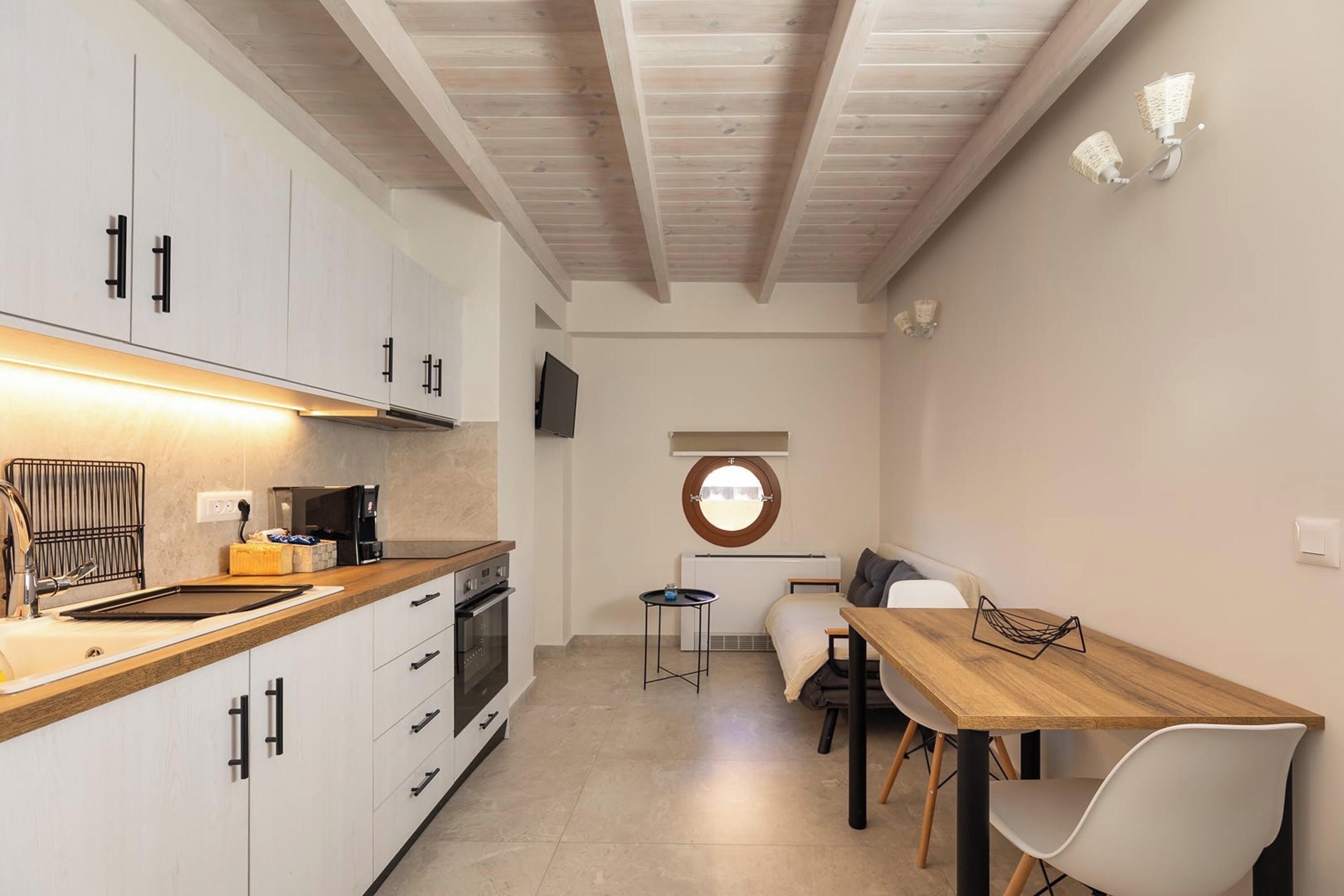 A compact kitchen with white cabinets, a wooden countertop, and a built-in oven. There is a dining area with a wooden table and white chairs. A small round window provides natural light.