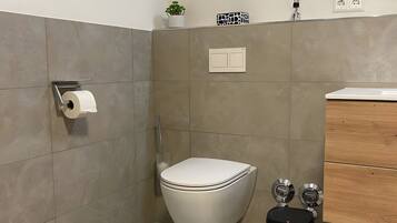 A bathroom with a toilet, bidet, and trash bin, tiled walls, and a small plant on the wall.