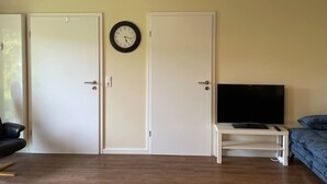 Una habitación con dos puertas blancas, un reloj de pared, un televisor de pantalla plana sobre un soporte blanco, un sofá azul y un suelo de madera.