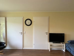 A room with two white doors, a wall clock, a flat-screen TV on a white stand, a blue sofa, and a wooden floor.
