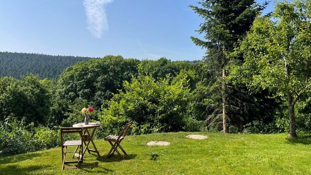 Un espacio al aire libre con césped, una mesa y sillas de madera, rodeado de árboles y una colina al fondo.