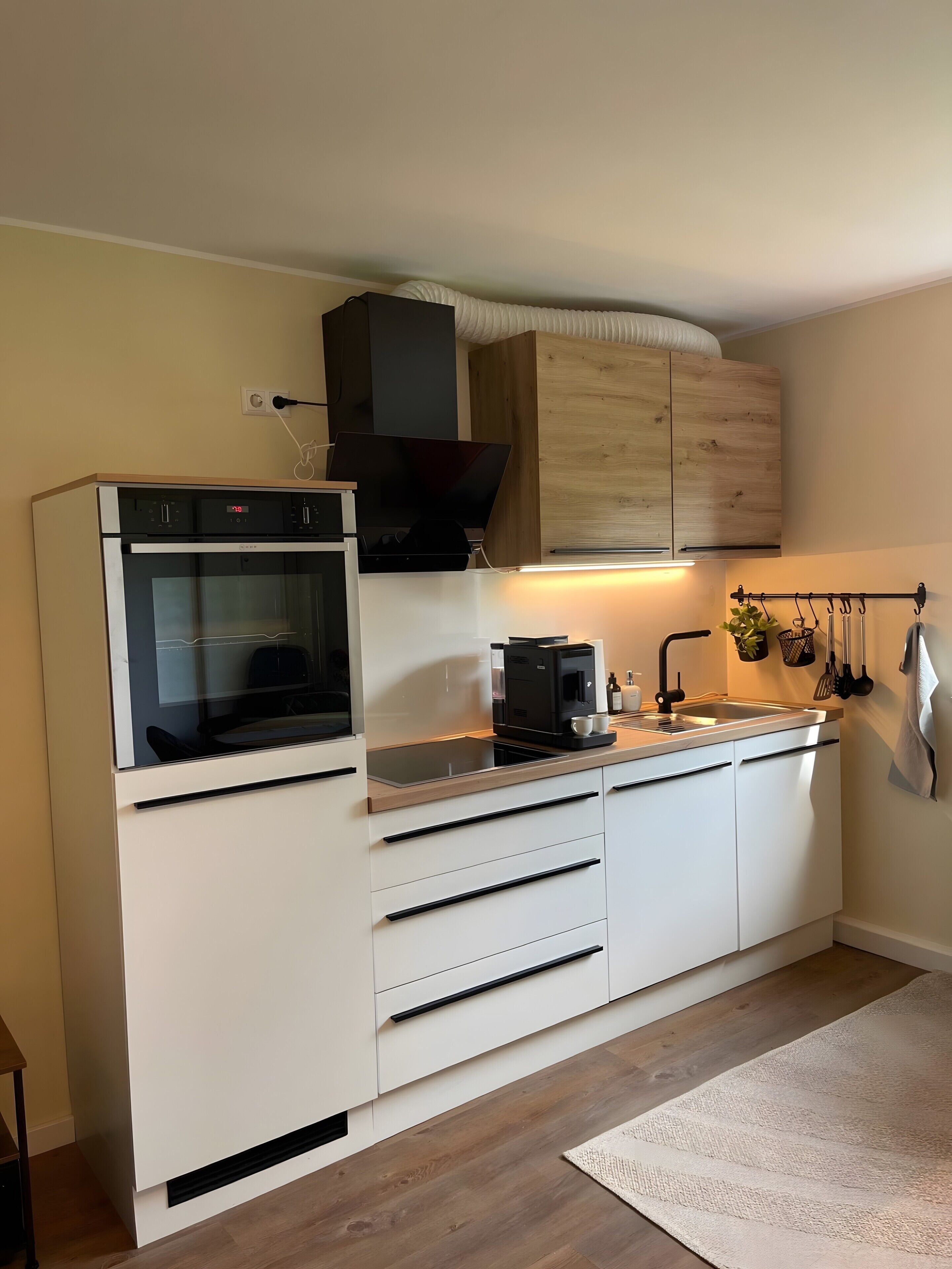 Een compacte keuken met een ingebouwde oven, een koffieapparaat en een houten kastje boven het aanrecht.