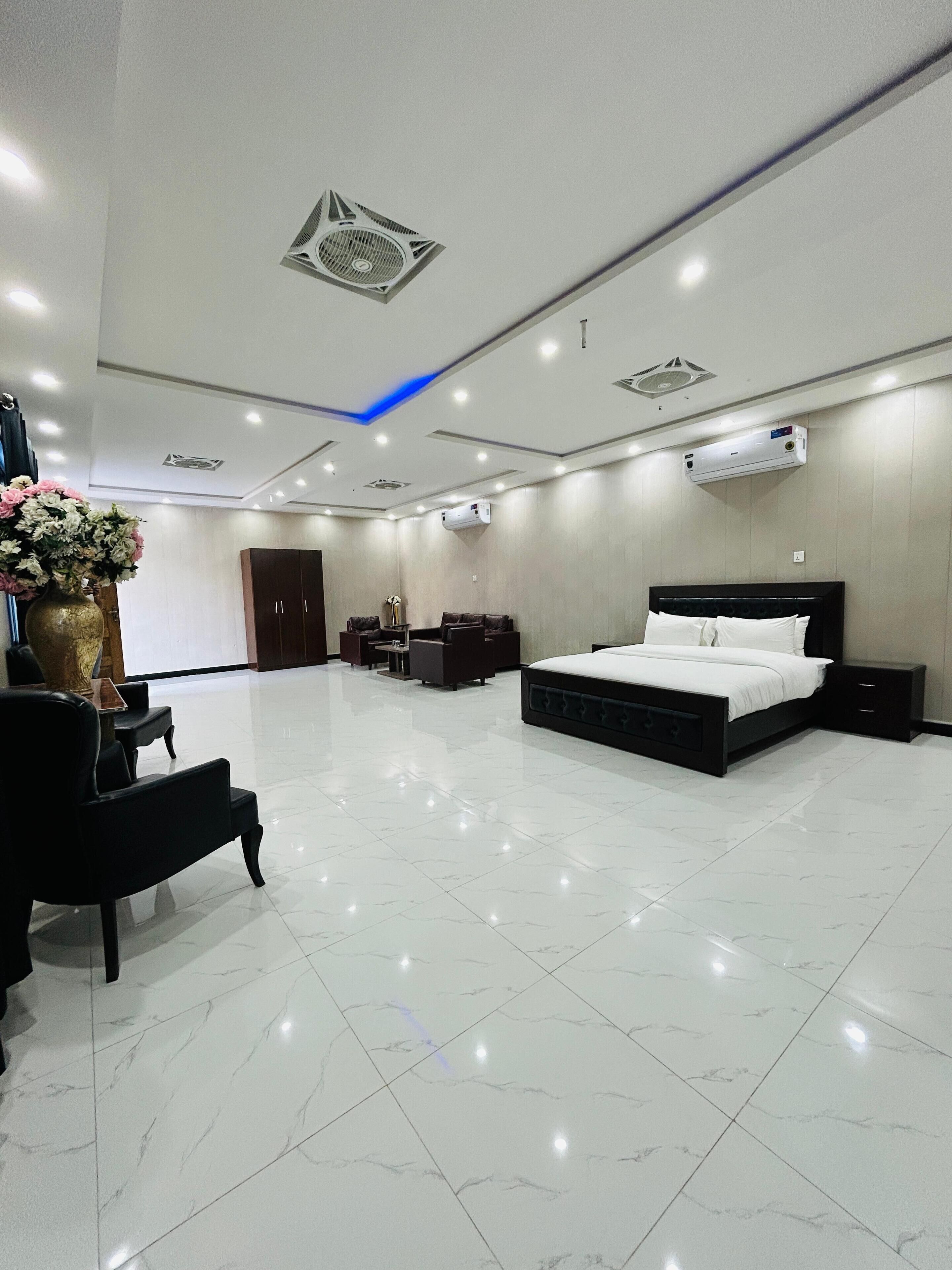 A modern hotel room with a large bed, a sitting area with a sofa and armchairs, a small table, and a decorative vase with flowers.
