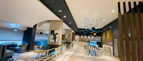 Un moderno vestíbulo de hotel con piso de patrón geométrico, un área de estar central con mesas y sillas, y una zona de bar con taburetes.