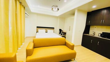 A hotel room with a bed, a yellow armchair, a desk, and a kitchenette.