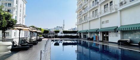 A swimming pool with a wooden deck and white pebbles, surrounded by white buildings with balconies.