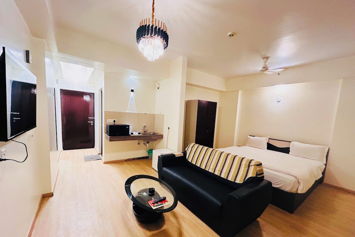 Habitación de hotel compacta con cama, sofá, una mesita con un control remoto, televisión y una kitchenette con microondas, además de un ventilador de techo.