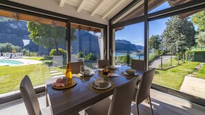 A dining area with a table set for six, large windows offering a view of a pool and mountains. - Villa Anna Lake View (Iseo)
