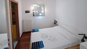 Een moderne slaapkamer met een houten vloer, een wit bed met blauw-witte streepkussens, een nachtkastje en een raam met decoratieve verlichting.