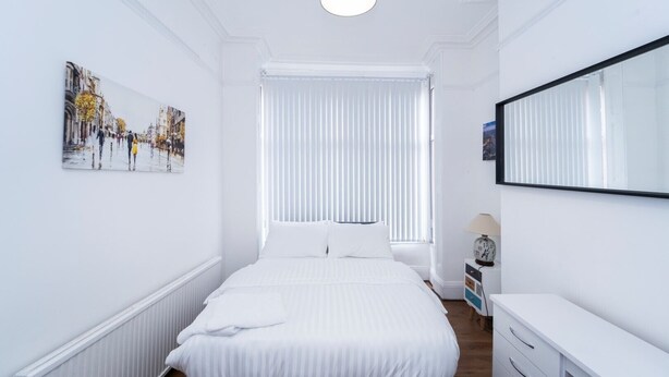 A modern bedroom with a large bed, white walls, a radiator, a window with blinds, and a painting on the wall.