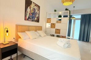 A neatly made bed with white linens and rolled towels, a bedside lamp, a wall-mounted painting, a white shelving unit with baskets, and a large window with curtains. -  Oceanografic Suite 1 (València)