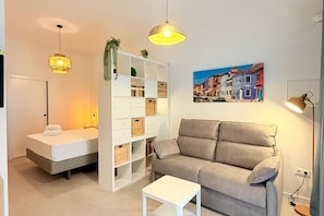 A modern living room with a sofa, a small white table, a bookshelf, and a painting of a European street scene. -  Oceanografic Suite 1 (València)