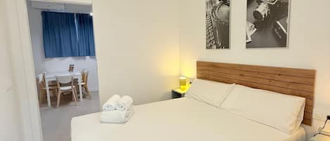 Camera d'albergo con un letto, un comodino con una lampada, un'area pranzo con tavolo e sedie, e due fotografie incorniciate appese al muro.