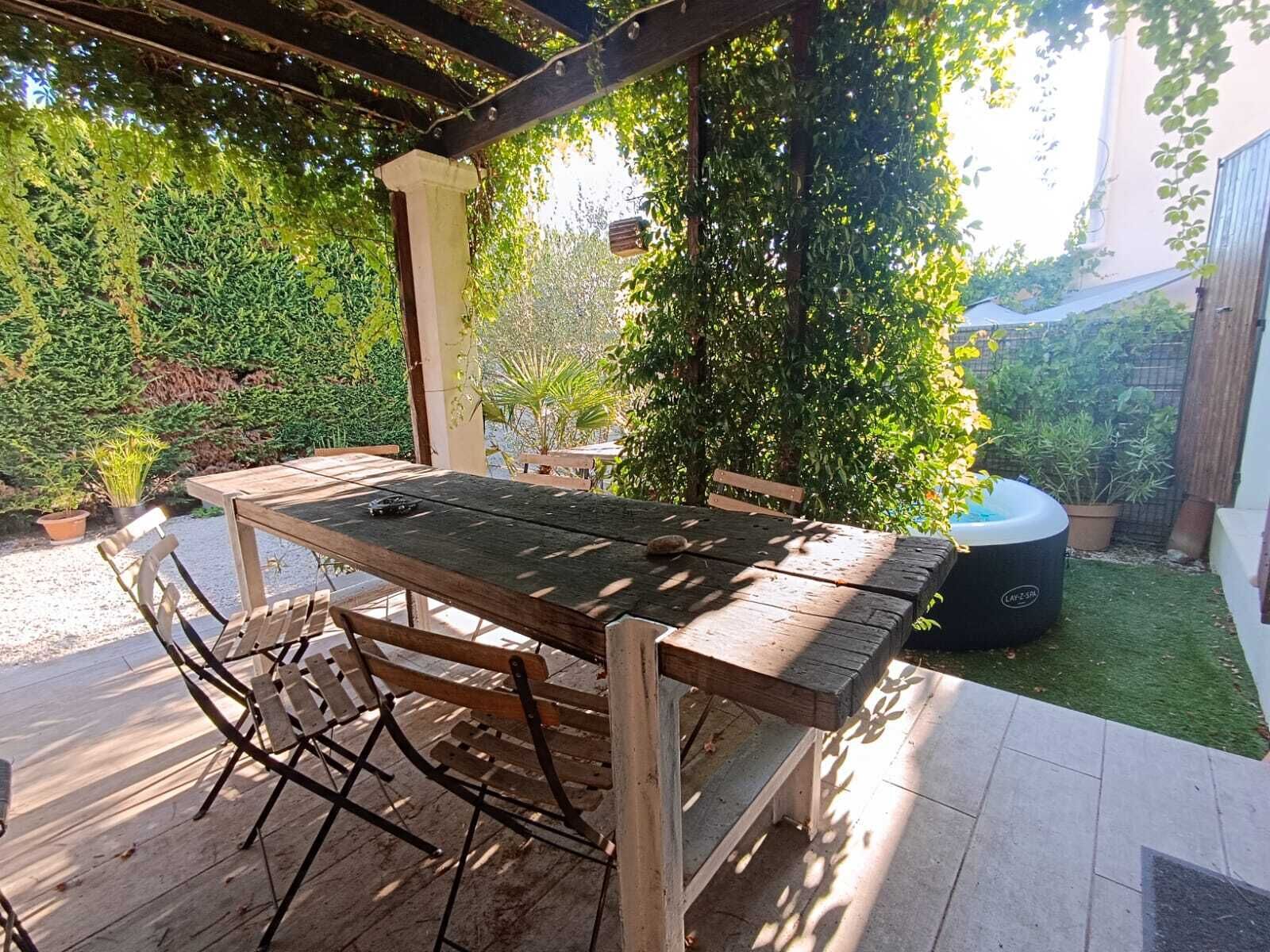 Une table et des chaises en bois, placées sous une pergola dans un jardin verdoyant, agrémenté d’un élément décoratif aquatique.