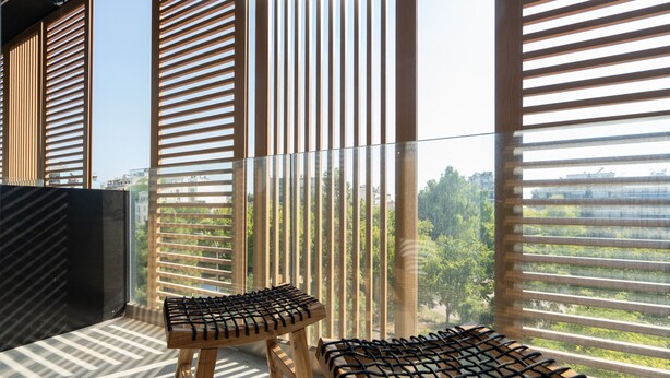 A balcony with two wooden stools and a glass railing, overlooking a cityscape and greenery. - LIV Athinum 1 - Luxury Apartment 3A (Athens)