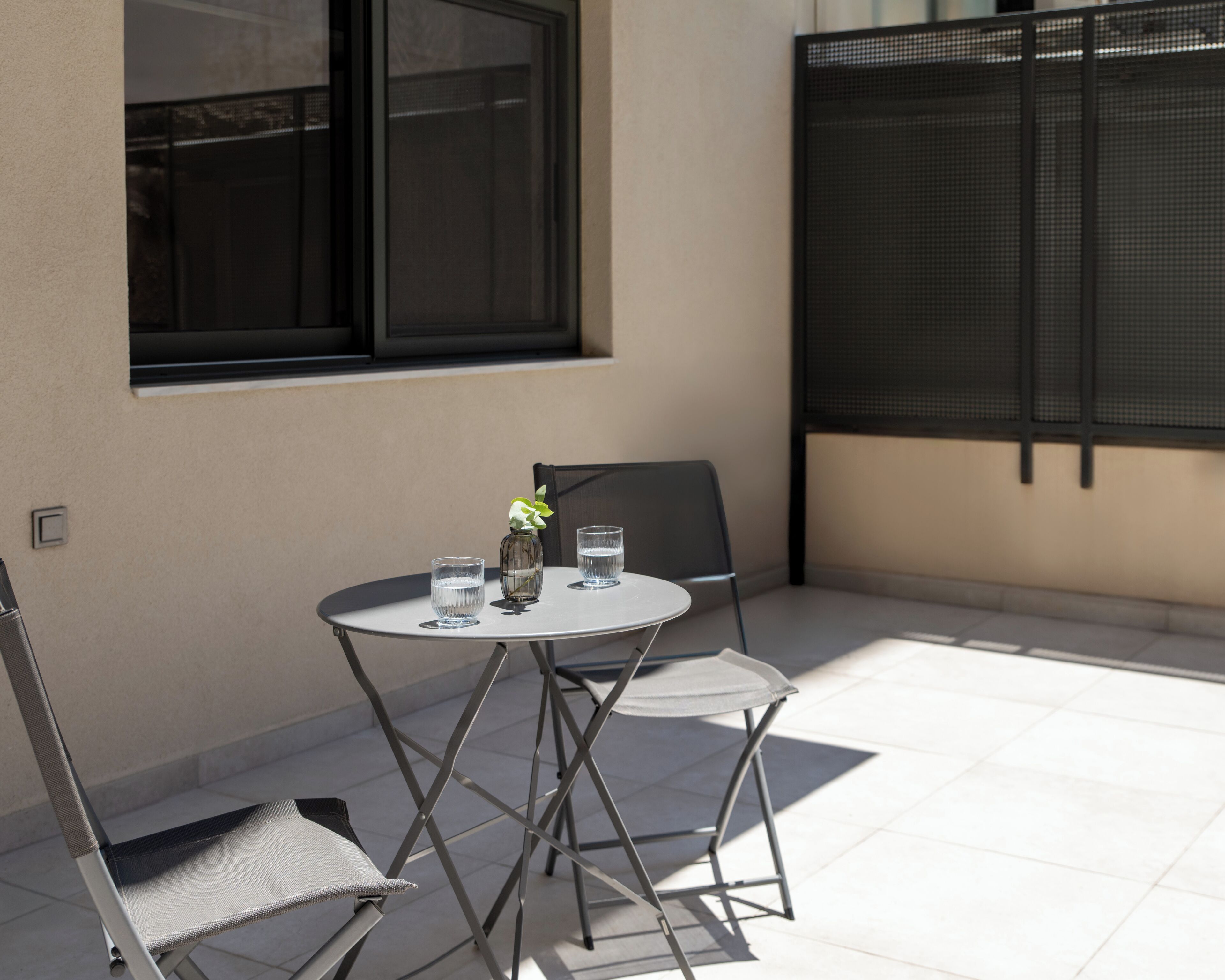 Un pequeño patio al aire libre con una mesa redonda y dos sillas, un florero con una flor y dos vasos de agua.