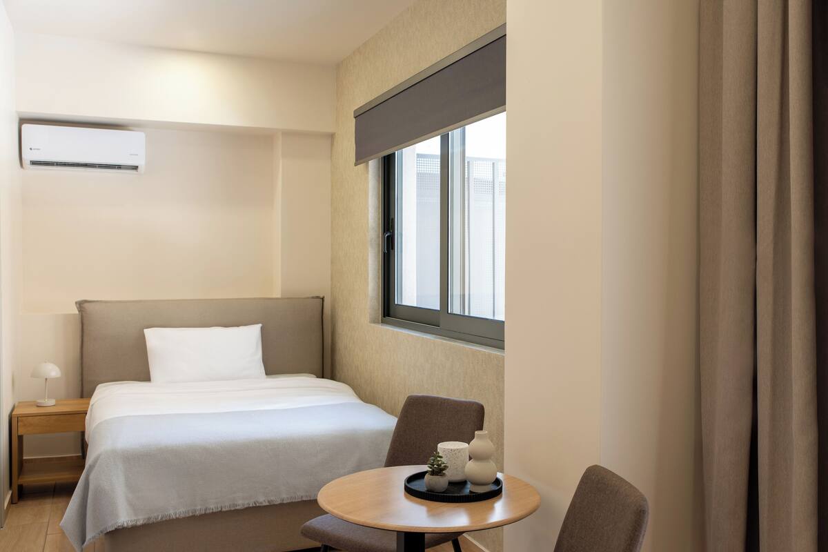Hotellihuone, jossa on sänky, yöpöytä, tuoli, pieni pöytä koriste-esineineen ja ikkuna, jossa on verhot.