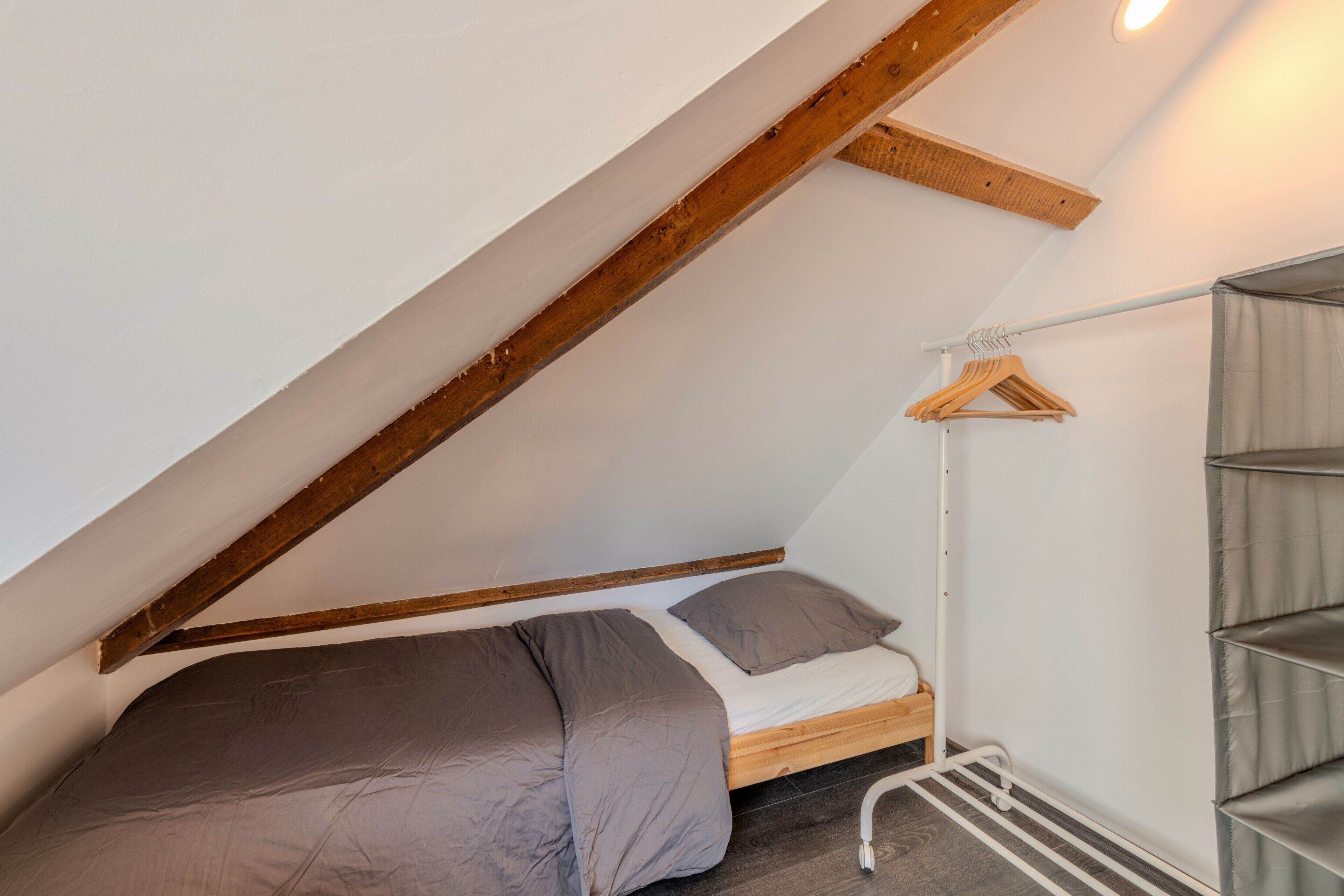 Ein kleiner Raum mit schrägen Wänden, einem Einzelbett, einem Holzkleiderbügel und einem Metallregal.