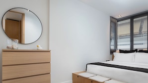 Un dormitorio moderno con un aparador de madera, un espejo redondo, una cama con ropa de cama blanca y una ventana con persianas.