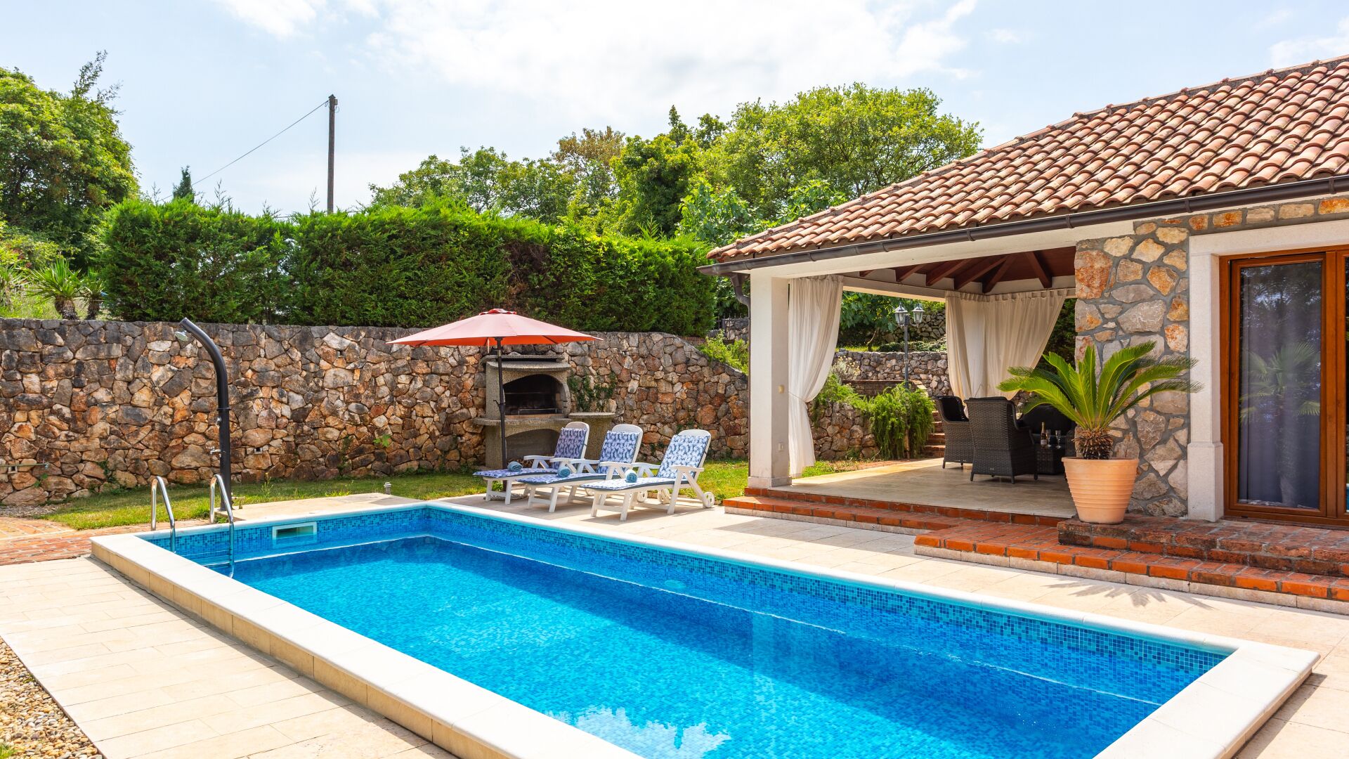A swimming pool with lounge chairs, a stone wall, and a covered patio area.