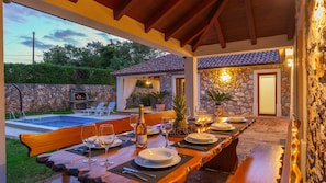 A covered outdoor dining area with a pool, stone walls, and a stone fireplace.
