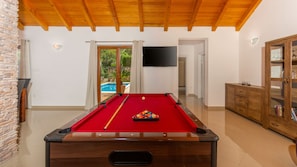 A pool table in a room with a wooden ceiling, a stone wall, and a view of an outdoor swimming pool.