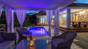 Un espace aménagé au bord de la piscine, avec des meubles en osier, une table où sont disposées des boissons, et un bâtiment en pierre en arrière-plan.