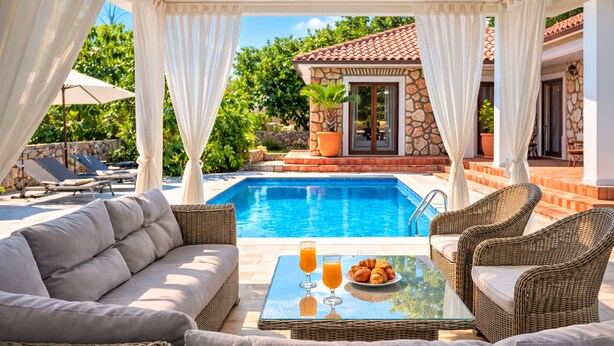 Un espace aménagé au bord de la piscine, avec un canapé, des fauteuils en osier, une table en verre sur laquelle sont disposés des boissons et des pâtisseries, et un bâtiment en pierre en arrière-plan.
