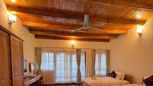 A room with wooden ceilings and floors, two beds with white linens, a desk with a chair, and a window with curtains.