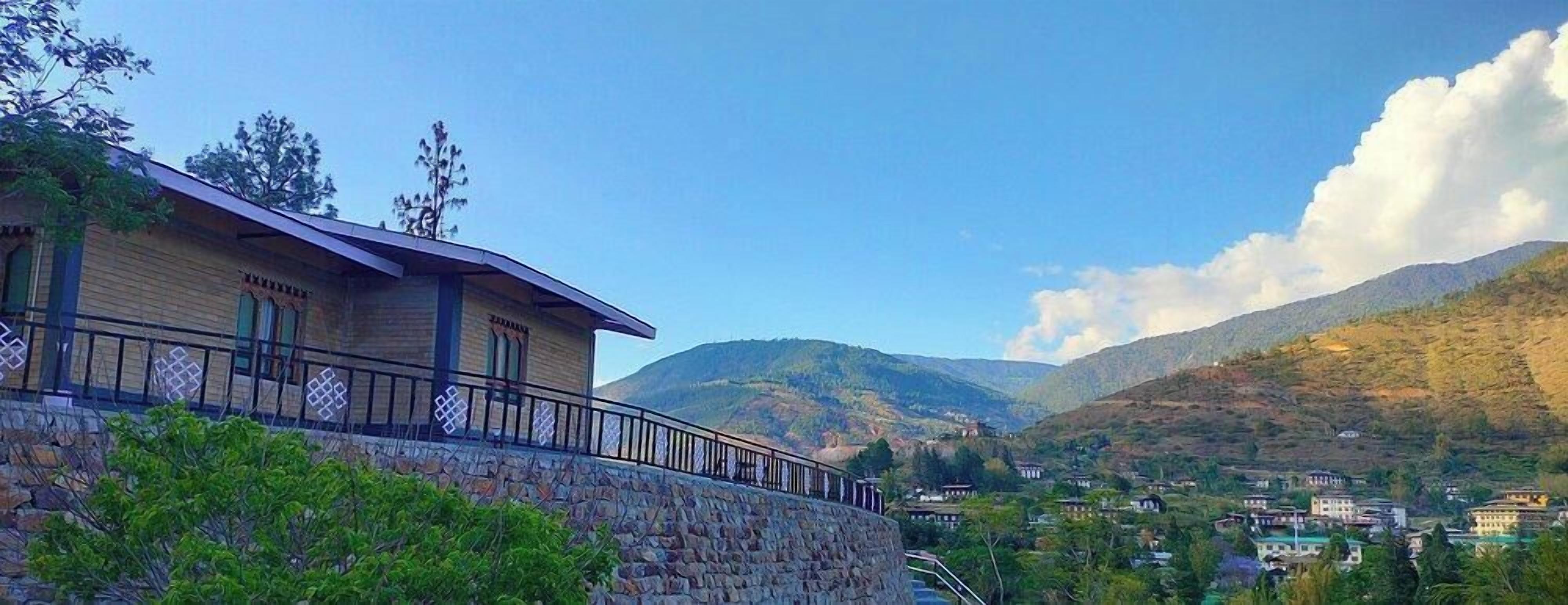 Edificio de paredes de piedra con un balcón, un paisaje montañoso al fondo y un pueblo abajo.