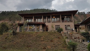 A two-story building with a flat roof and multiple windows, surrounded by a hilly landscape.