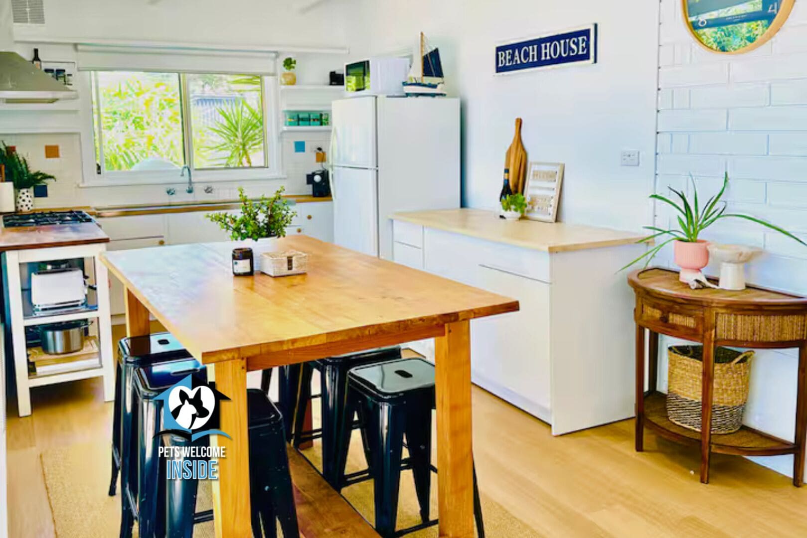 A modern kitchen with a wooden table, bar stools, and a sign reading 'BEACH HOUSE' on the wall.