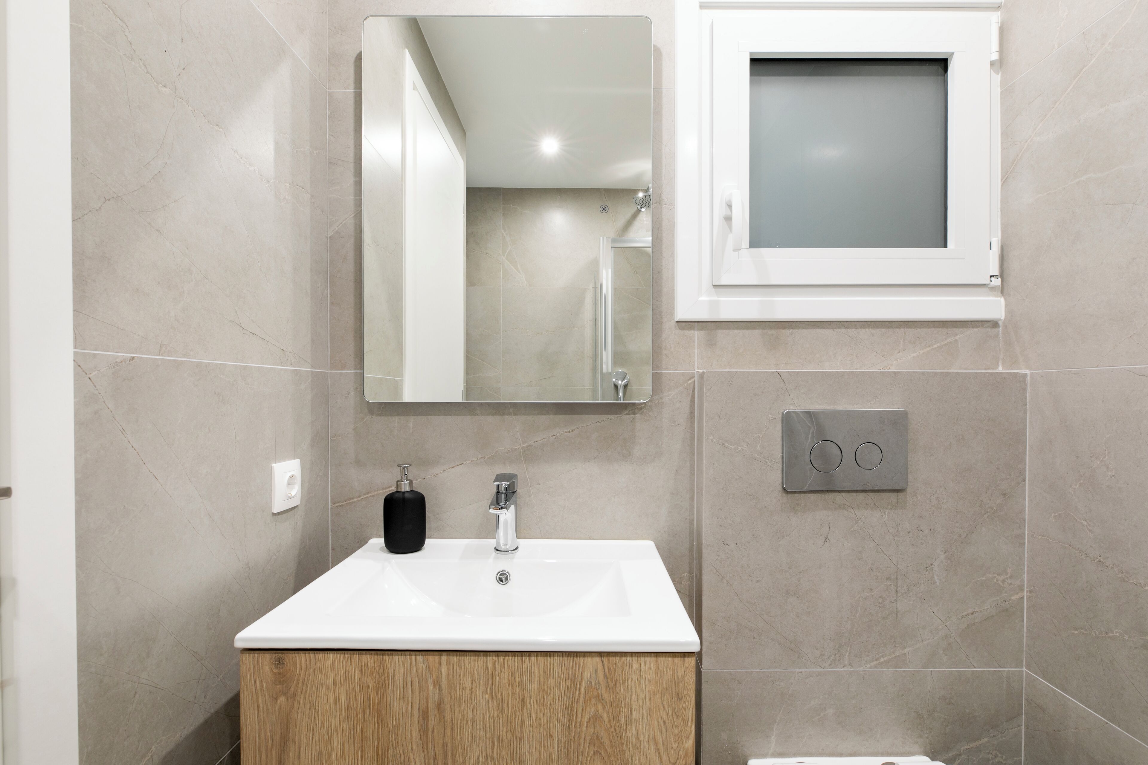 Un baño con lavabo blanco, tocador de madera, espejo y ventana con cristal esmerilado.