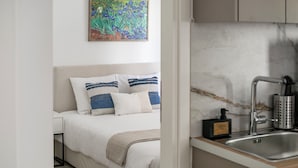 A bedroom with a bed, pillows, a nightstand, a painting on the wall, and a kitchen area with a sink and utensils. - Anatoli - Athenian 2-BR Apartment in Athens (Zografos)