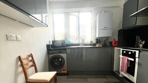 A compact kitchen with a washing machine, a microwave, and a stove. There is a chair and a window providing natural light. - London Luxe Apartment - Spacious & Stylish (London)